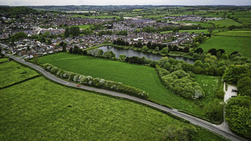 Aerial view of green fields and trees beside a winding rural road, with a large pond and surrounding houses forming the edge of a village in the background.