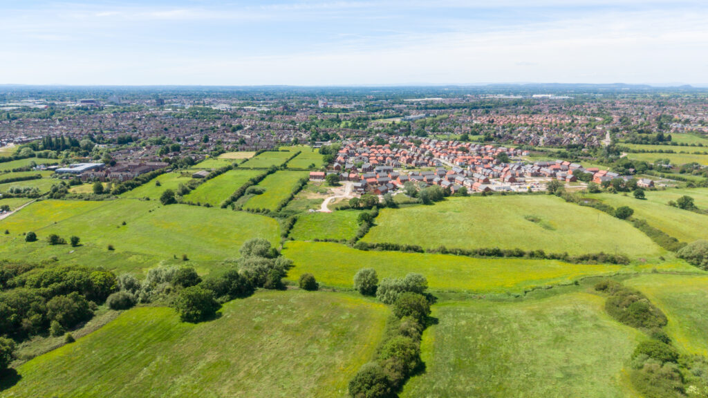 Aerial view over hedged grassland towards a new-build housing estate under construction on the edge of Crewe, with the wider town stretching beyond.