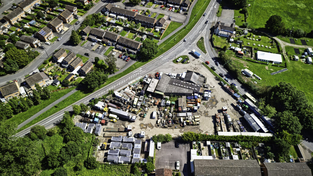 Overhead view of a brownfield commercial yard of workshops, plant, vehicles and portable cabins, set in the fork of a road junction beside terraced housing.