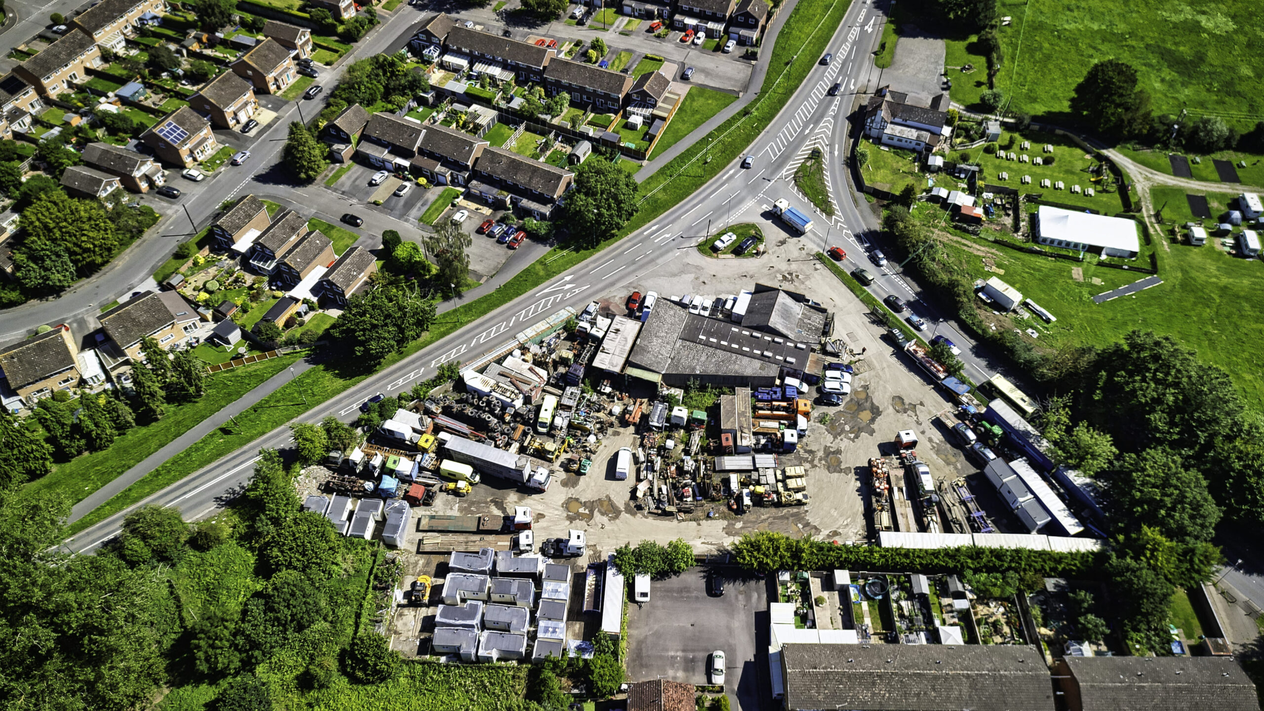 Overhead view of a brownfield commercial yard of workshops, plant, vehicles and portable cabins, set in the fork of a road junction beside terraced housing.